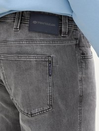 Grijze denim jeans met een zwart leren label met de tekst "TOM TAILOR." Zichtbare achterzak met een klein merkembleem. Zachte textuur en standaardontwerp.