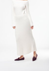 Abito bianco in maglia a costine con spalle scoperte, maniche lunghe e lunghezza midi, abbinato a scarpe basse viola scuro con un accento decorativo.