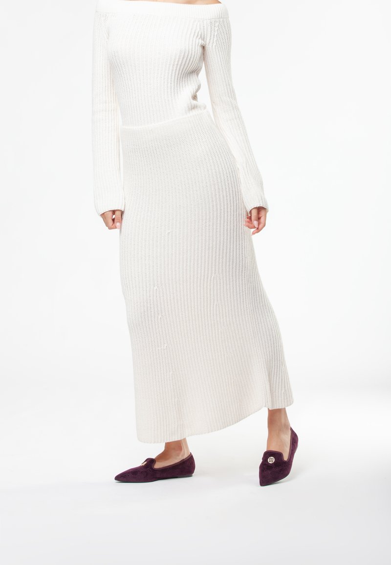 Abito bianco in maglia a costine con spalle scoperte, maniche lunghe e lunghezza midi, abbinato a scarpe basse viola scuro con un accento decorativo.
