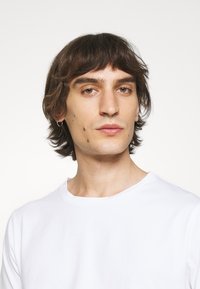 Tricou alb cu guler rotund. Țesătura pare netedă și moale. Părul este de lungime medie și de culoare închisă, cu o unduire subtilă.