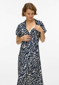Femme aux cheveux courts portant une robe à motifs bleu marine et blanc, tenant l'encolure ouverte pour révéler un sous-vêtement blanc.