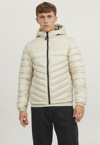Padded jacket in cream, featuring a hood, black zip closure, and chevron stitch design. Worn with dark jeans, contrasting color details.