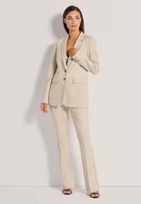 Beige op maat gemaakt pak met een enkelvoudige blazer met twee knopen en recht gesneden broek. Gladde stof met een lichte glans.