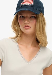 Lichtgrijs geribbelde t-shirt met korte mouwen, voorzien van een V-hals versierd met delicate kanten rand. Marineblauwe honkbalpet met een ontwerp van de Amerikaanse vlag.
