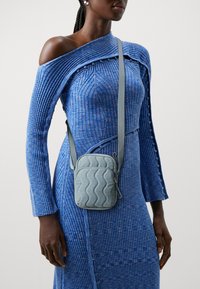 Prešívaná crossbody taška v svetlomodrej farbe s vlnkovaným vzorom. Obsahuje zips a nastaviteľný popruh. Nosí sa cez modré textúrované šaty s odhalenými ramenami.