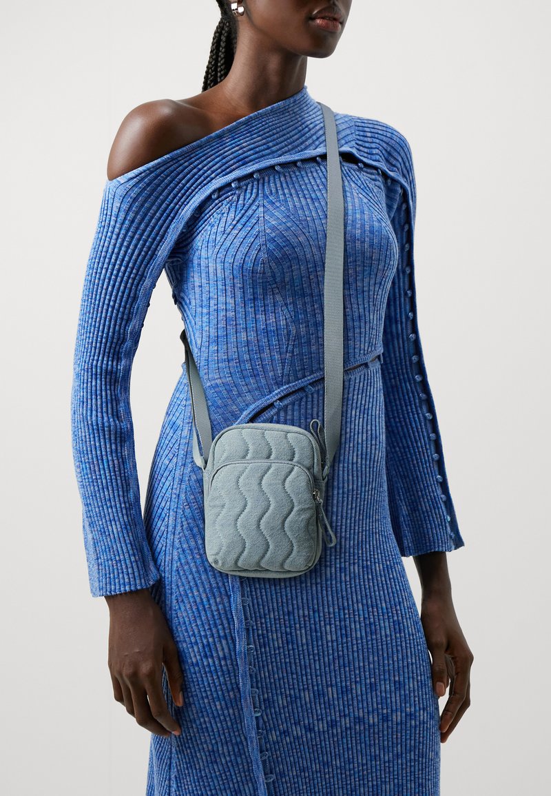 Prešívaná crossbody taška v svetlomodrej farbe s vlnkovaným vzorom. Obsahuje zips a nastaviteľný popruh. Nosí sa cez modré textúrované šaty s odhalenými ramenami.