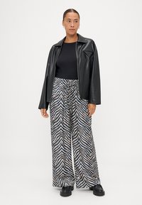 Jaqueta de couro sintético preta, blusa justa preta e calças largas com padrão de riscas de zebra em bege e azul-marinho. Calçado robusto preto.