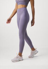 Leggings alti in vita di colore viola, realizzati in un tessuto elasticizzato, con un design aderente e un discreto dettaglio del logo. Indossati con scarpe da ginnastica di colore rosa chiaro.