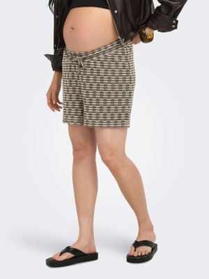 Personne enceinte portant une jupe à motifs, un top court noir, une veste en cuir marron et des sandales noires, debout avec une main sur la hanche.
