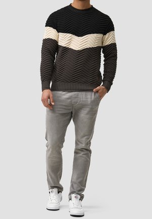 Pull en tricot avec un motif zigzag texturé en noir, crème et gris foncé. Associé à un pantalon gris clair et des baskets blanches.