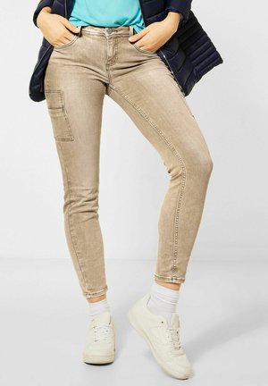 Personne portant un pantalon cargo beige slim, des baskets blanches, des chaussettes blanches, une chemise bleue et une veste noire avec les mains dans les poches.