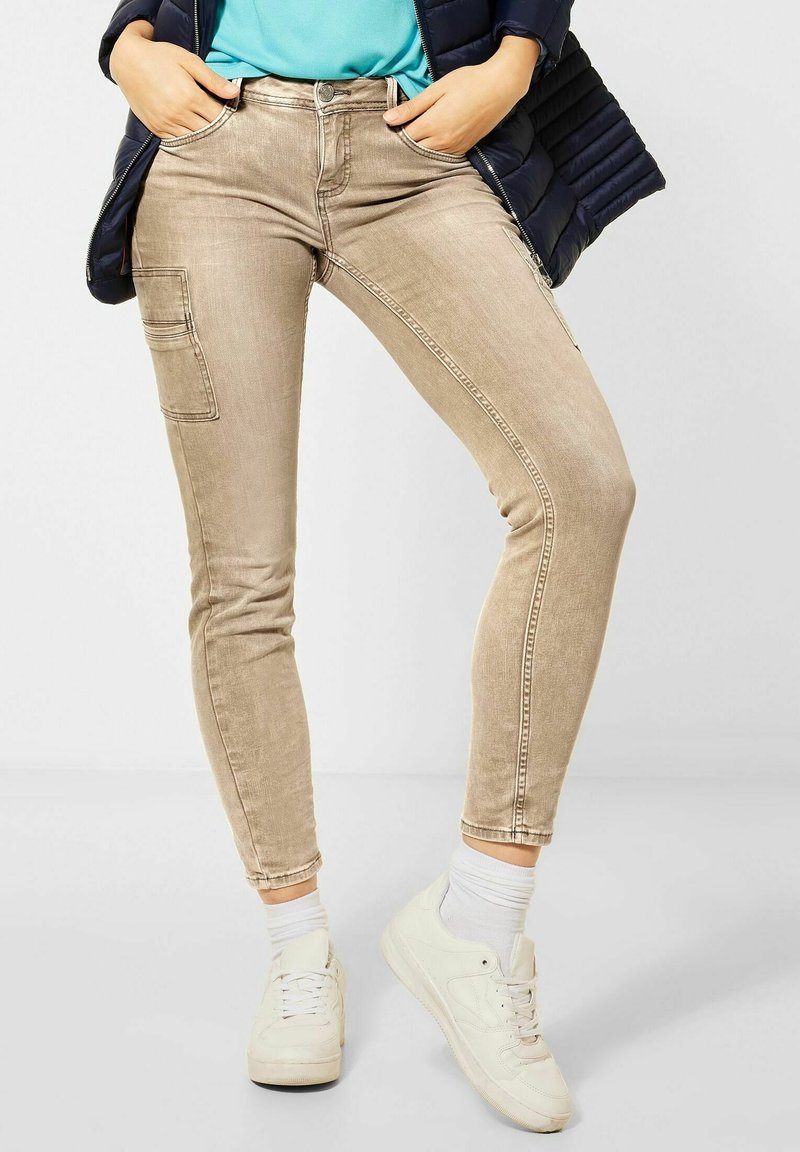 Personne portant un pantalon cargo beige slim, des baskets blanches, des chaussettes blanches, une chemise bleue et une veste noire avec les mains dans les poches.