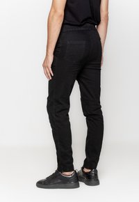 Czarne dżinsy w wąskim kroju, z elastycznym paskiem i dwiema tylnymi kieszeniami. Połączone z czarnymi trampkami na neutralnym tle.