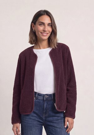 Femme aux cheveux bruns mi-longs, portant une chemise blanche, une veste polaire zippée violet foncé et un jean bleu, souriant à la caméra.