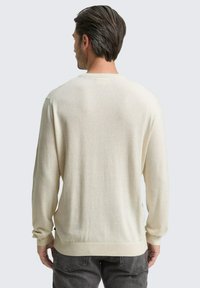 Mann mit braunen Haaren, der einen cremefarbenen langärmligen Pullover und dunkelgraue Jeans trägt, steht mit dem Rücken zu einer einfarbigen grauen Hintergrund.