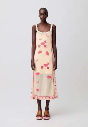 Mujer con vestido crema sin mangas con bordado floral rosa y sandalias marrones de plataforma, de pie frente a un fondo claro y liso.