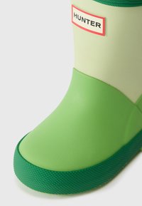 Botte de pluie en caoutchouc verte avec une semelle texturée, dotée d'un dessus vert clair et d'une étiquette de logo "HUNTER" rouge et blanche.