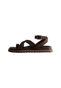 Sandal con cinturino incrociato marrone, dotata di fibbia, con una soletta testurizzata e una suola piatta in gomma. Il design enfatizza il comfort e la semplicità.