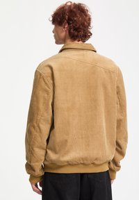Veste en velours côtelé marron avec des rayures texturées, col boutonné et taille élastique. Elle possède des manches longues avec des poignets côtelés.