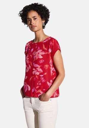 Blusa rossa a fiori con maniche corte, caratterizzata da motivi rosa e viola; tessuto leggero con vestibilità comoda, abbinata a pantaloni bianchi.