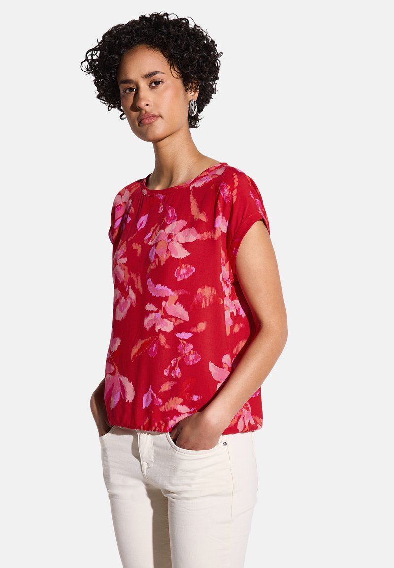 Blusa rossa a fiori con maniche corte, caratterizzata da motivi rosa e viola; tessuto leggero con vestibilità comoda, abbinata a pantaloni bianchi.