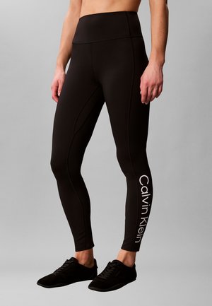 Vrouw draagt zwarte leggings met hoge taille met "Calvin Klein" logo op het onderbeen en zwarte sneakers, staand tegen een grijze achtergrond.