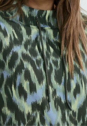 Gros plan sur une personne portant un sweat à capuche à motifs verts et bleus avec des cordons noirs et de longs cheveux bruns partiellement visibles.