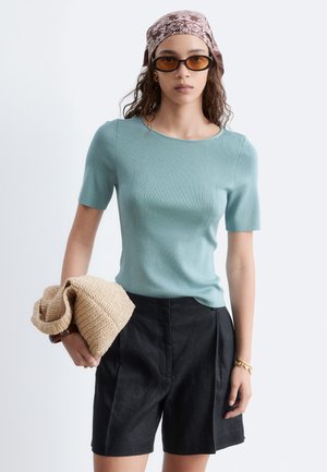 Femme portant un haut bleu clair à manches courtes, un short noir taille haute, un foulard marron à motifs, des lunettes de soleil orange, et tenant un pull en maille beige.