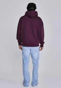 Přetažená vínová mikina s velkou kapucí, žebrovanými manžetami a lemem. Kombinována se světle modrými džíny se rovnými nohavicemi. Hladká, pohodlná tkanina.