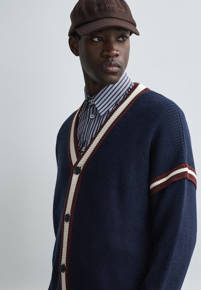 Cardigan de malha azul marinho com decote em V, com riscas brancas e bordô em contraste nas mangas, e botões pretos na parte da frente.