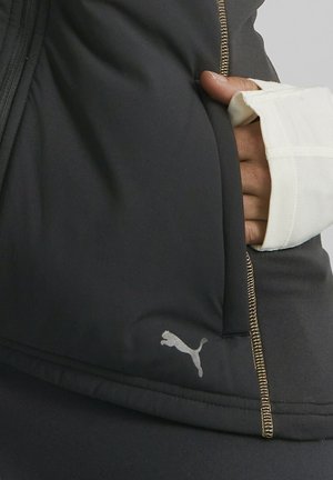Mano en manga blanca con orificio para el pulgar, sosteniendo una chaqueta negra de Puma con el logo de Puma visible y detalle de cremallera con costuras doradas.