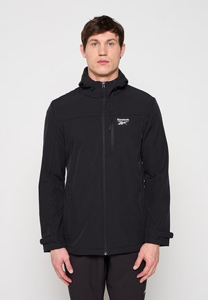 Mężczyzna w czarnej, zapinanej na zamek kurtce z kapturem marki Reebok, stojący prosto na jednolitym jasnym tle.