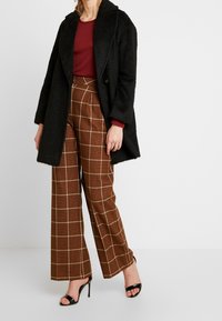 Svart texturerad kappa över en burgundy topp, matchad med bruna rutiga vida byxor och svarta remsandaler.