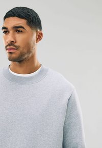 Sudadera gris con una textura suave, cuello acanalado y costuras de hombro ligeramente caídas. Llevada sobre una camiseta blanca, plano cercano de la parte superior del cuerpo.