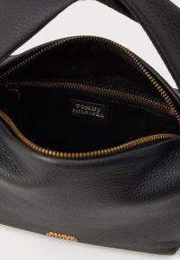 Černá kožená kabelka s texturovaným povrchem, zlatým zipem a štítkem s nápisem "TOMMY HILFIGER" uvnitř přihrádky.