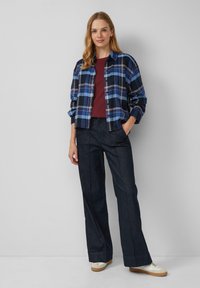 Chemise à carreaux bleu marine et bleu, associée à un haut bordeaux et un jean large foncé. Le mannequin porte des baskets blanches avec des accents beiges.