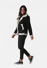 Veste en peau lainée noire avec doublure blanche, fermeture éclair et détail ceinture. Associée à un jean noir et des baskets, le mannequin est posé de profil.
