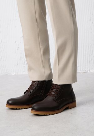 Brązowe skórzane botki za kostkę o gładkim wykończeniu, okrągłym nosku i kontrastowej, jasnobrązowej podeszwie. Udoskonalone brązowymi sznurówkami i detalami w postaci przyszycia.