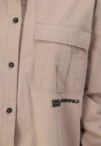 Beige színű, könnyű anyagból készült ing, mellzsebbel és fekete gombokkal. A "Karl Lagerfeld Jeans" fekete hímzéssel van feltüntetve.