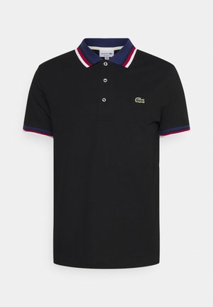 Homme portant un polo noir avec un col rouge, blanc et bleu, un pantalon cargo beige, des baskets bleues, tenant une veste matelassée couleur beige dans la main gauche.