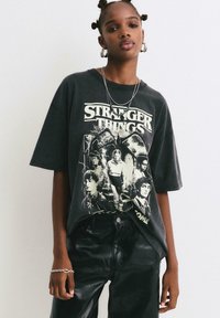 Crni oversized majica s grafičkim printom "Stranger Things" u bijeloj boji, kratki rukavi, pamučni materijal, uparen s crnim kožnim hlačama.
