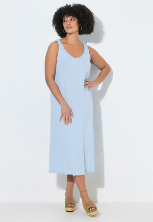 Donna dai capelli ricci che indossa un vestito midi azzurro chiaro senza maniche e sandali platform color cuoio, in piedi su sfondo bianco.