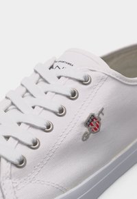 Λευκό πάνινο αθλητικό παπούτσι με λευκά κορδόνια, μεταλλικές τρύπες και μικρό κεντημένο λογότυπο με ασπίδα και γράμματα στο πλάι.