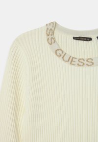 Maglione a coste color crema con scollo rotondo, caratterizzato da un logo "GUESS" testurizzato a contrasto sul collo. Tessuto a maglia morbido e elasticizzato.