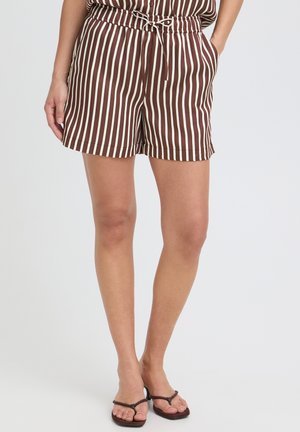 Žena nosi hnedé a bielo pruhované šortky s vertikálnymi pruhmi, ktoré majú šnúrku v páse, a tmavé pletené sandále, stojaca na jednoduchom pozadí.