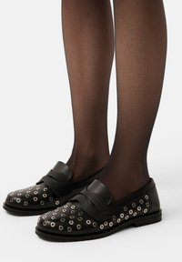 Mocassins noirs avec œillets en métal, détail de sangle texturé et tissu lisse. Portés avec des collants noirs transparents, mettant en avant un design discret.