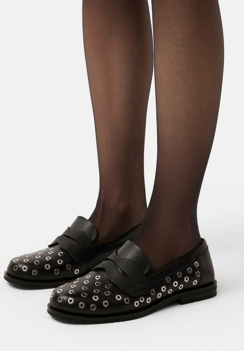 Mocassins noirs avec œillets en métal, détail de sangle texturé et tissu lisse. Portés avec des collants noirs transparents, mettant en avant un design discret.