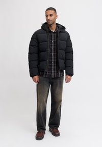 Chaqueta de plumas negra con capucha, diseño acolchado, cierre de cremallera, llevada sobre una camisa de cuadros. Combinada con jeans negros desgastados y zapatos marrones.