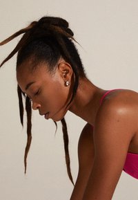 Haut de bikini texturé rose, cheveux lisses avec des dreadlocks, boucles d'oreilles en argent, arrière-plan atténué mettant en avant le profil et le teint de la silhouette.