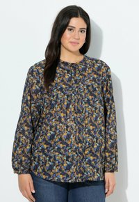 Frau mit langen dunklen Haaren, die eine blau, orange und schwarz gemusterte Bluse mit Knöpfen und blaue Jeans trägt, steht vor einer weißen Wand.